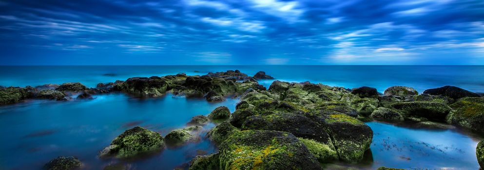 Dusk (Sunset) over green moss covered black volcanic rocky outcrop in the middle of the blue ocean with calm sea and sky full of rolling cloud in Port Fairy, Victoria, Australia.