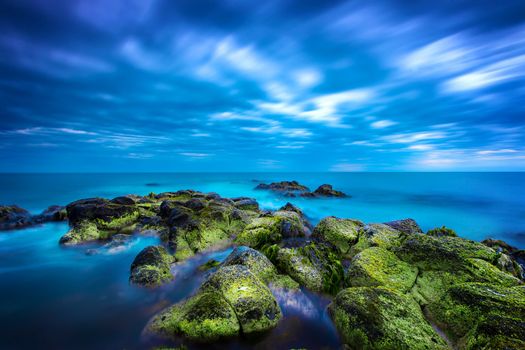 Dusk (Sunset) over green moss covered black volcanic rocky outcrop in the middle of the blue ocean with calm sea and sky full of rolling cloud in Port Fairy, Victoria, Australia.