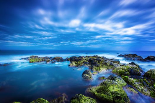 Dusk (Sunset) over green moss covered black volcanic rocky outcrop in the middle of the blue ocean with calm sea and sky full of rolling cloud in Port Fairy, Victoria, Australia.