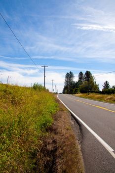 A rural highway travels up and over a hill with trees and blue sky.