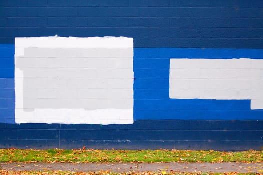 A dark blue wall of a building next to a street with autumn leaves during the fall that has grey rectangles of paint to cover up graffiti.