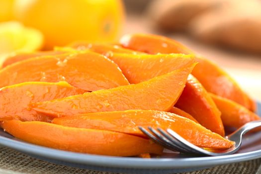 Sweet potato wedges caramelized with brown sugar and fresh orange juice with a fork on plate, oranges and sweet potatoes in the back (Selective Focus, Focus one third into the wedges)