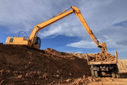 Heavy excavator loading dumper truck with sand in quarry over blue sky