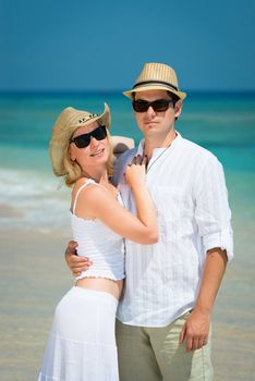 Embracing young couple in sunglasses and white dress on a tropical beach with blue sea on background