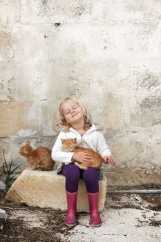 A little preschool child holding a cat, with her eyes closed, sitting against a wall on a farm