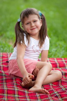 Little smiling cute blond girl preschooler with ponytails sitting on the red plaid on green grass in summer