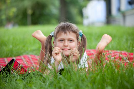 Portrait of little cute blond girl preschooler with ponytails who is lying on red plaid in park with green grass summer