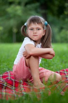 Portrait of little militant blond girl preschooler with ponytails sitting in closed pose on the red plaid on green grass in summer