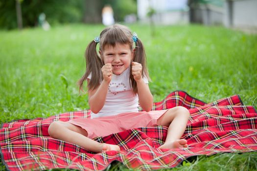 Little angry cute blond girl preschooler with ponytails sitting on the red plaid on green grass in summer
