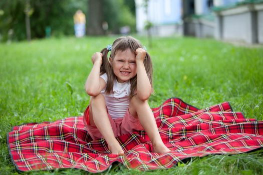 Little angry cute blond girl preschooler with ponytails sitting on the red plaid on green grass in summer