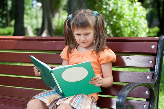 Nice little studious girl preschooler is reading open book and sitting on the red plaid on green grass in summer park