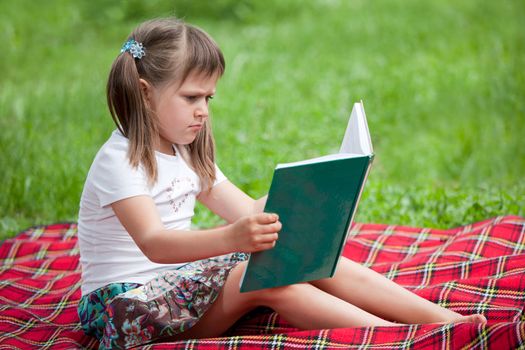 Little studious girl preschooler is reading open book and sitting on the red plaid on green grass in summer park