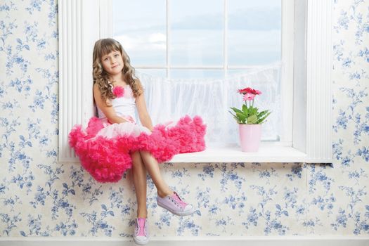 Romantic dreaming young girl dressed luxuriant wavy ball dress with frill sitting on white windowsill inside room near fresh natural flower in flowerpot