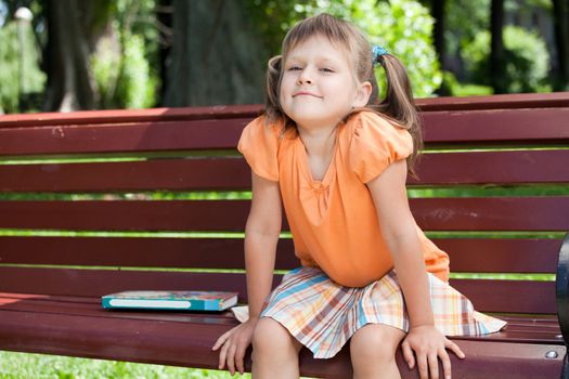 Little cute smiling girl preschooler with open book who is sitting on the wooden bench in summer park