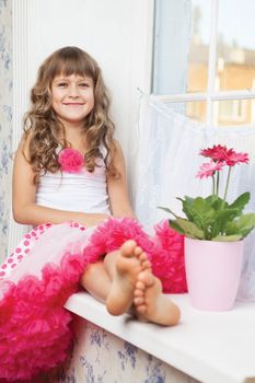 Young amazing joyful girl teenager close-up dressed luxuriant wavy skirt with frill sitting with outstretched legs on white windowsill inside room near fresh natural flower in flowerpot