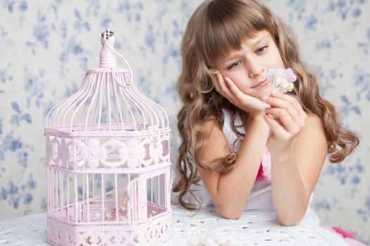 Portrait of tender sincere dreamy romantic openhearted blond girl with long wavy hair looking at flower seating near open empty pink birdcage and lacy white table on the light blue flower background