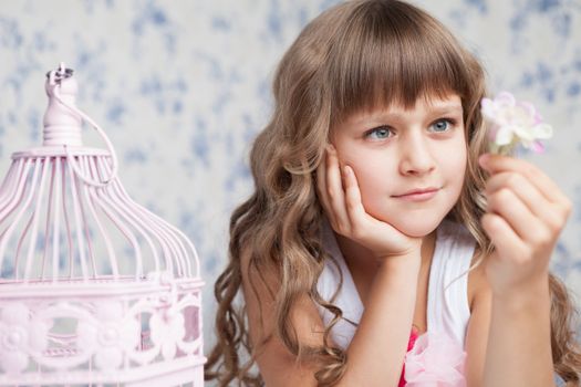 Portrait of tender sincere dreamy romantic openhearted blond girl with long wavy hair looking at pink flower in hand near birdcage on the light blue flower background