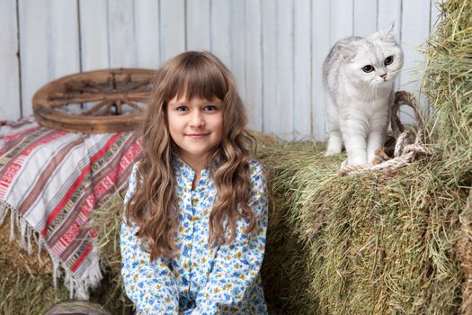 Portrait of sincere little blond girl villager and white cat on hay stack in wooden hayloft