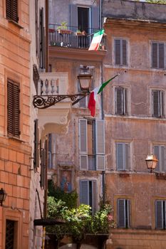 Picturesque facades of old medieval multistory urban residential stone brick-red houses with flowers and Italian flags on balconies, with street lamp and icon on wall in city Rome Italy