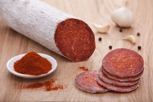 Traditional Hungarian speciality sliced sausage salami on wooden board with garlic, corns of black pepper and bowl with milled paprika spice