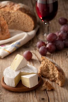 Piece and wheel of traditional Normandy soft cheese Camembert with country homemade bread, grapes and glass of red wine on wooden table