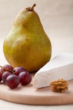 Close-up ripe green pear, piece of white soft cheese brie, core of Circassian walnut and red grapes on wooden board and linen tablecloth