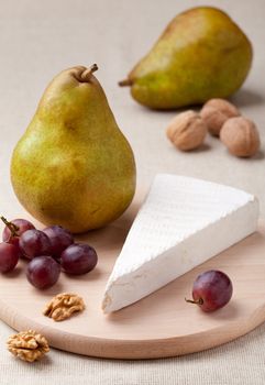 Ripe green pears, piece of white soft cheese brie, cores of Circassian walnuts and red grapes on wooden board and linen tablecloth