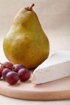 Close-up ripe green pear, piece of white soft cheese brie, red grapes on wooden board and linen tablecloth