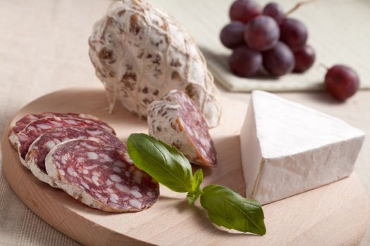 Close-up Traditional sliced meat sausage salami on wooden board with triangular soft cheese brie, green basil and bunch of grapes on textile towel