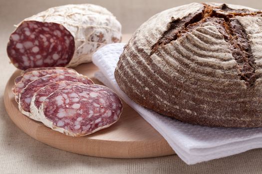 Close-up traditional sliced meat sausage salami on wooden board with loaf of homemade brown bread  on textile towel