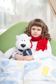 Sympathetic little sick girl with red scarf embraces white toy bear under blanket in bed