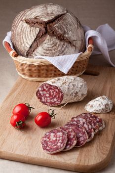 Traditional sliced salami on wooden board, fresh cherry tomatoes brown bread loaf in wicker breadbasket
