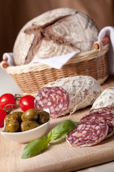 Traditional sliced salami on wooden board, fresh cherry tomatoes, green olives in salad bowl, brown bread loaf in wicker breadbasket