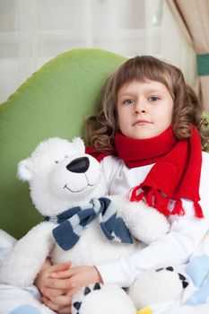 Sympathetic little sick girl with red scarf embraces white toy bear under blanket in bed