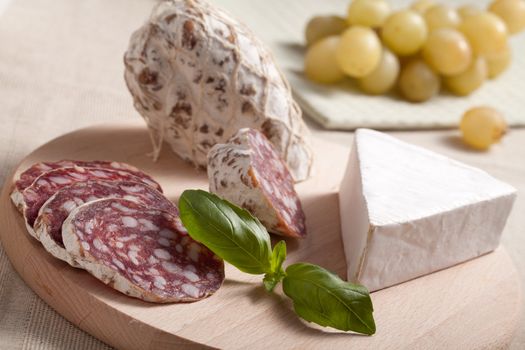 Close-up Traditional sliced meat sausage salami on wooden board with triangular soft cheese brie, green basil and bunch of grapes on textile towel