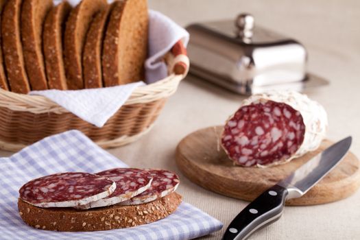 Served kitchen table, sandwich on napkin, salami on wooden board and knife, metal butterdish, sliced brown bread in wicker breadbasket