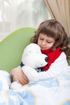 Sympathetic little sick girl with red scarf embraces white toy bear under blanket in bed