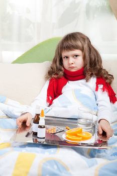 Sympathetic little sick girl in red scarf under blanket in bed is taking a salver with pills, sprays and orange