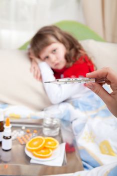 Close-up clinical thermometer in woman’s hand showing fever and little sick girl wrapped in red scarf under blanket in the bed and tray with pills, sprays, oranges and glass of water