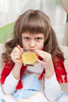 Close-up sympathetic little sick girl wrapped in red scarf under blanket in bed is drinking antiviral with lemon slice from transparent cup