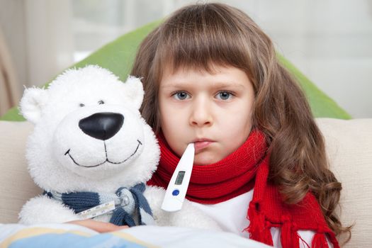 Sympathetic little sick girl with red scarf and clinical thermometer in mouth embraces white toy bear under blanket in the bed