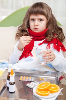 Sympathetic little sick girl wrapped in red scarf is taking a pill and a salver with pills, sprays and orange on the blanket in the bed