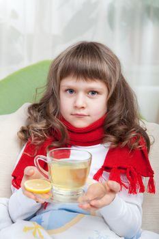 Close-up sympathetic little sick girl wrapped in red scarf under blanket in bed is drinking antiviral with lemon slice from transparent cup