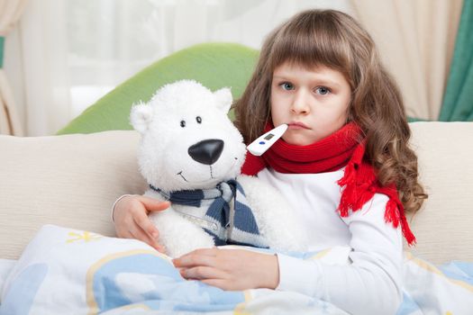 Sympathetic little sick girl with red scarf and clinical thermometer in mouth embraces white toy bear under blanket in the bed