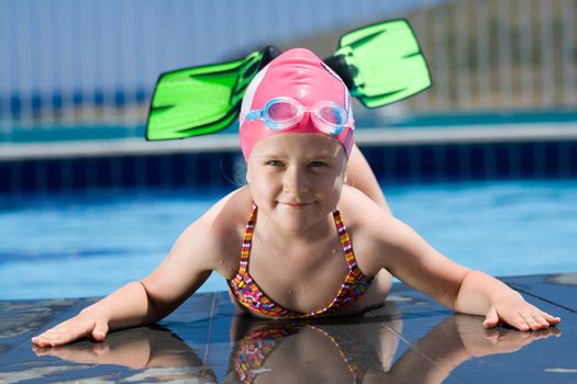 Close-up portrait of smiling cute little child in bathing cap, glasses and fins is lying on the swimming pool ledge