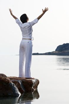 Young dressed white woman with raised arms meets sunrise staying barefoot on the sea stone. Back lighting