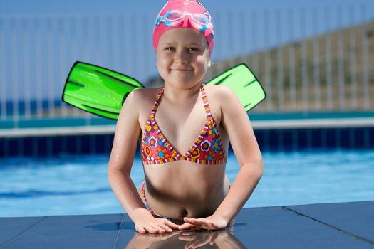 Close-up portrait of smiling cute little child in bathing cap, glasses and fins is lying on the swimming pool ledge