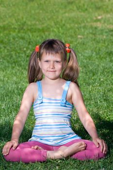 Smiling happy little cute girl who is sitting and meditating in yoga lotus asana under tree shadow on the green grass