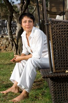 Young barefoot relaxing woman dressed white suit sits on wicker park bench