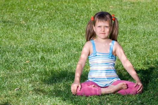 Cute little girl is sitting in yoga lotus asana under tree shadow on the green grass